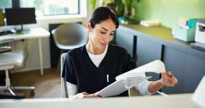 A woman in dark scrubs sits at a modern desk, looking down and reviewing papers. Her dark hair is pulled back with a pen in her pocket. Office furnishings, a plant, and sunlight through a window are behind her.