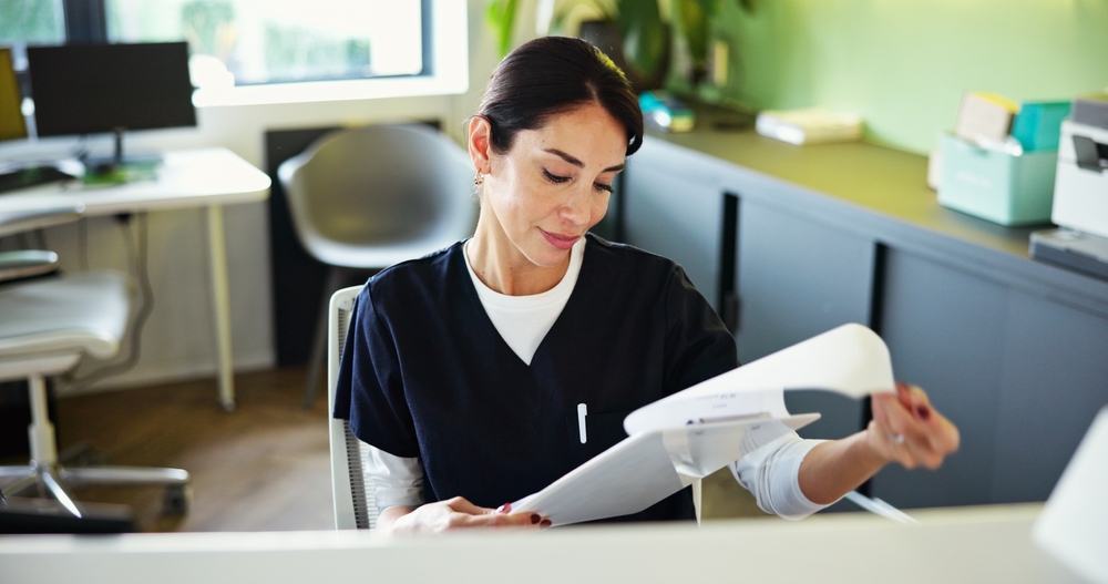 A woman in dark scrubs sits at a modern desk, looking down and reviewing papers. Her dark hair is pulled back with a pen in her pocket. Office furnishings, a plant, and sunlight through a window are behind her.