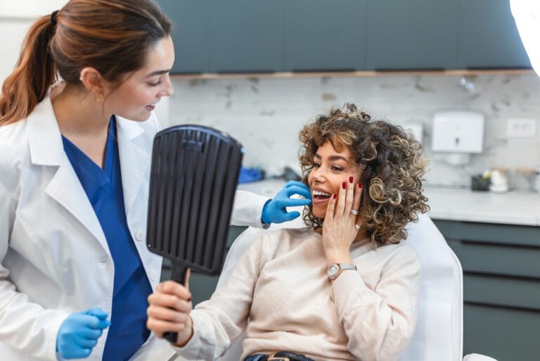 A smiling woman with curly hair holds a mirror and touches her cheek while sitting in a dental chair. A dentist in a white coat and blue gloves talks to her; both look happy in a modern clinic.