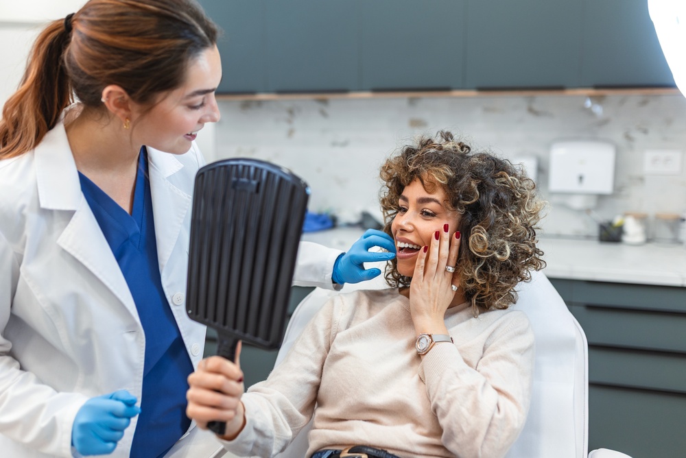A smiling woman with curly hair holds a mirror and touches her cheek while sitting in a dental chair. A dentist in a white coat and blue gloves talks to her; both look happy in a modern clinic.